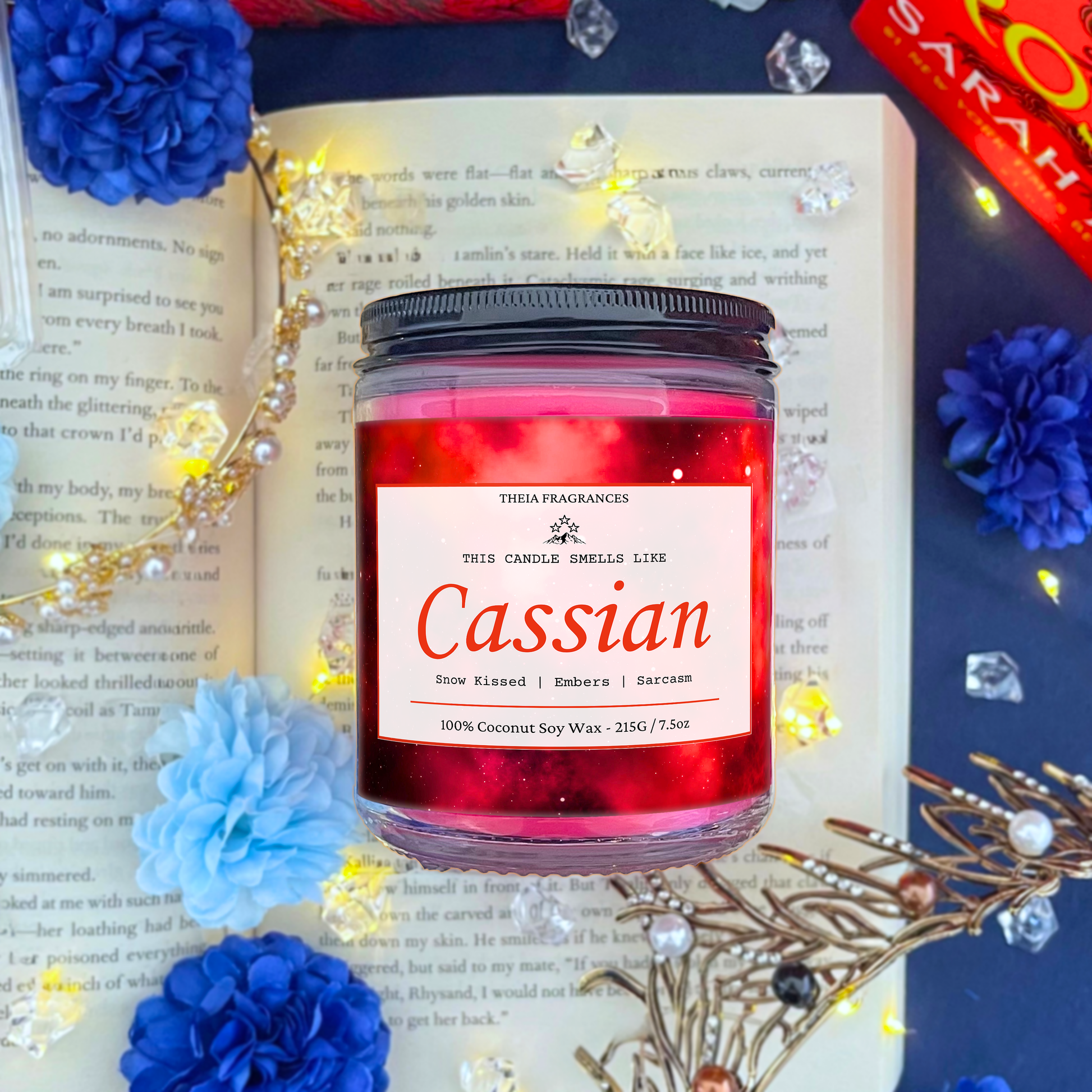 The 'Sarcastic Illyrian Warrior' coconut soy wax candle by Theia Fragrances is demonstrated in the 215G glass jar with a black lid. The label features a red galaxy. It is styled on an open book with decorative crystals and flowers.