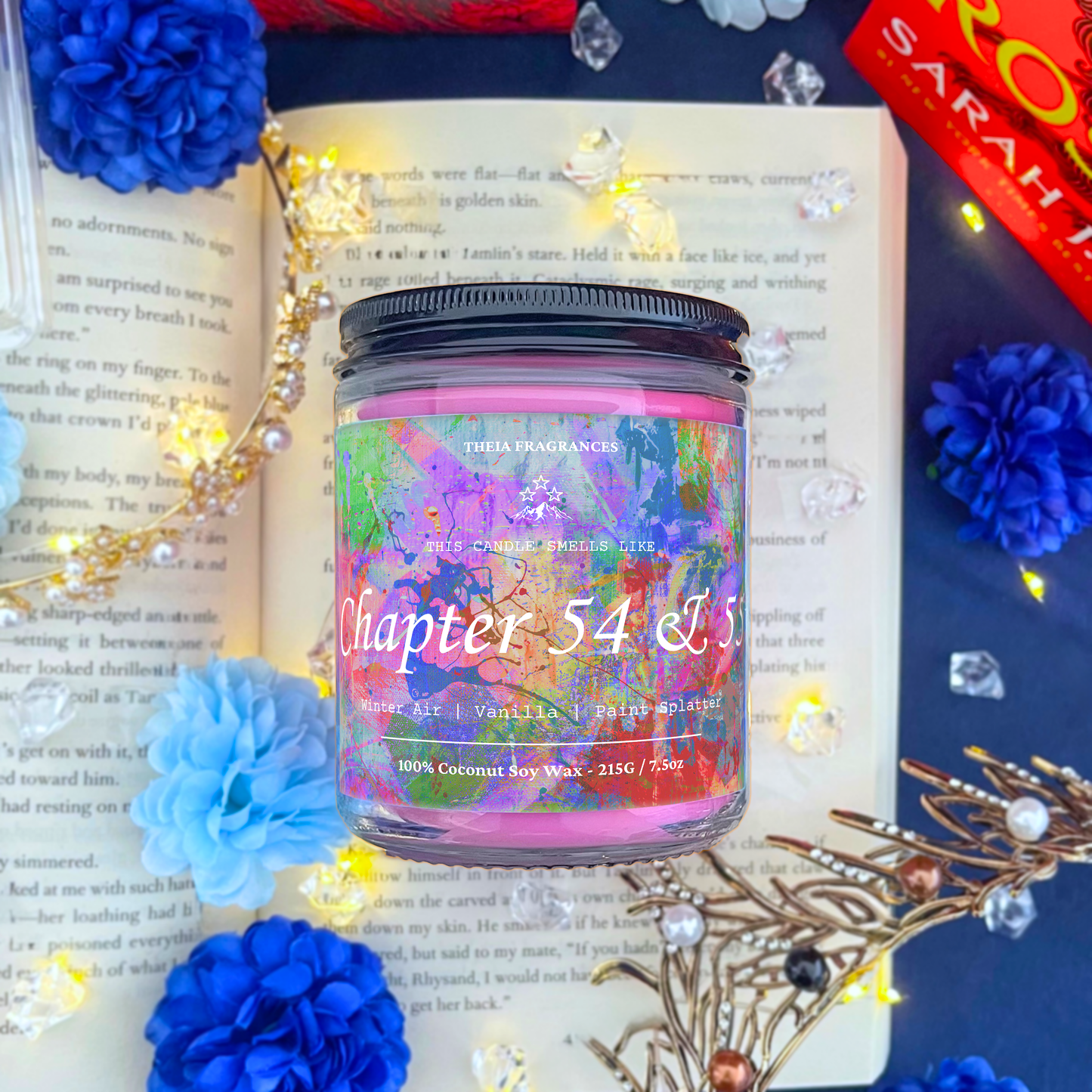 The 'Chapter 54 & 55' coconut soy wax candle by Theia Fragrances is demonstrated in the 215G glass jar with a black lid. The label features a paint splattered background. It is styled on an open book with decorative crystals and flowers.