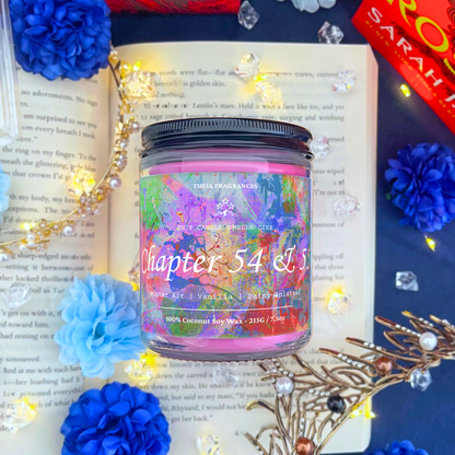 The 'Chapter 54 & 55' coconut soy wax candle by Theia Fragrances is demonstrated in the 215G glass jar with a black lid. The label features a paint splattered background. It is styled on an open book with decorative crystals and flowers.