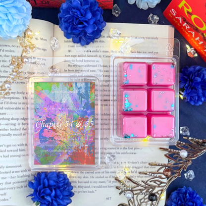 The 'Chapter 54 & 55' coconut soy wax candle by Theia Fragrances is demonstrated in the 78G rectangular wax melt. The label features a paint splattered background. It is styled on an open book with decorative crystals and flowers.
