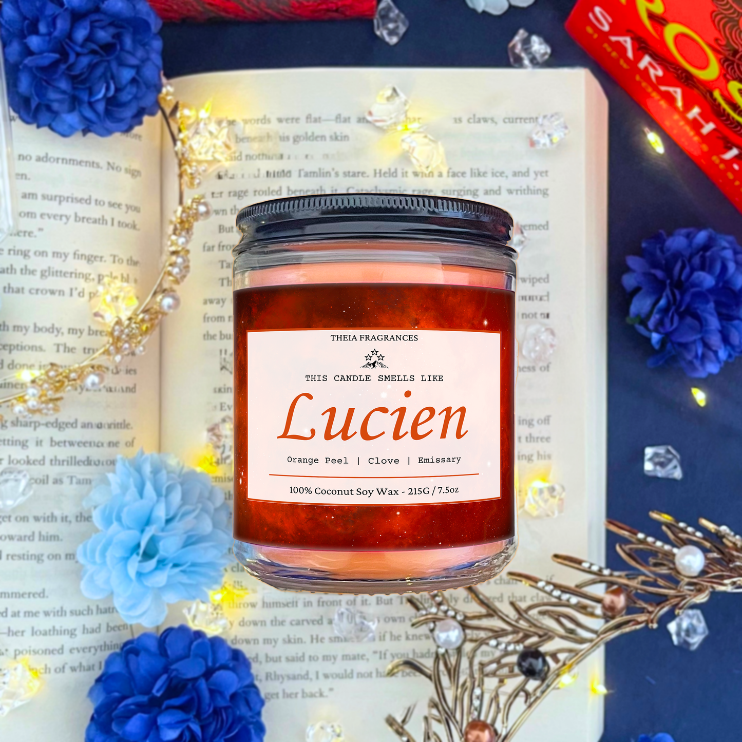 The 'Emissary' coconut soy wax candle by Theia Fragrances is demonstrated in the 215G glass jar with a black lid. The label features an orange galaxy. It is styled on an open book with decorative crystals and flowers.