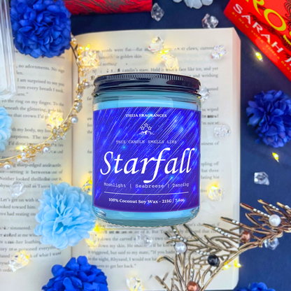The 'Starfall' coconut soy wax candle by Theia Fragrances is demonstrated in the 215G glass jar with black lid. The label features a blue starry sky. It is styled on an open book with decorative crystals and flowers.