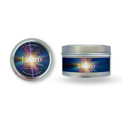 The 'Velaris' coconut soy wax candle by Theia Fragrances is demonstrated in the 11G tin candle. The label features a burst of starlight. It is styled on an open book with decorative crystals and flowers.