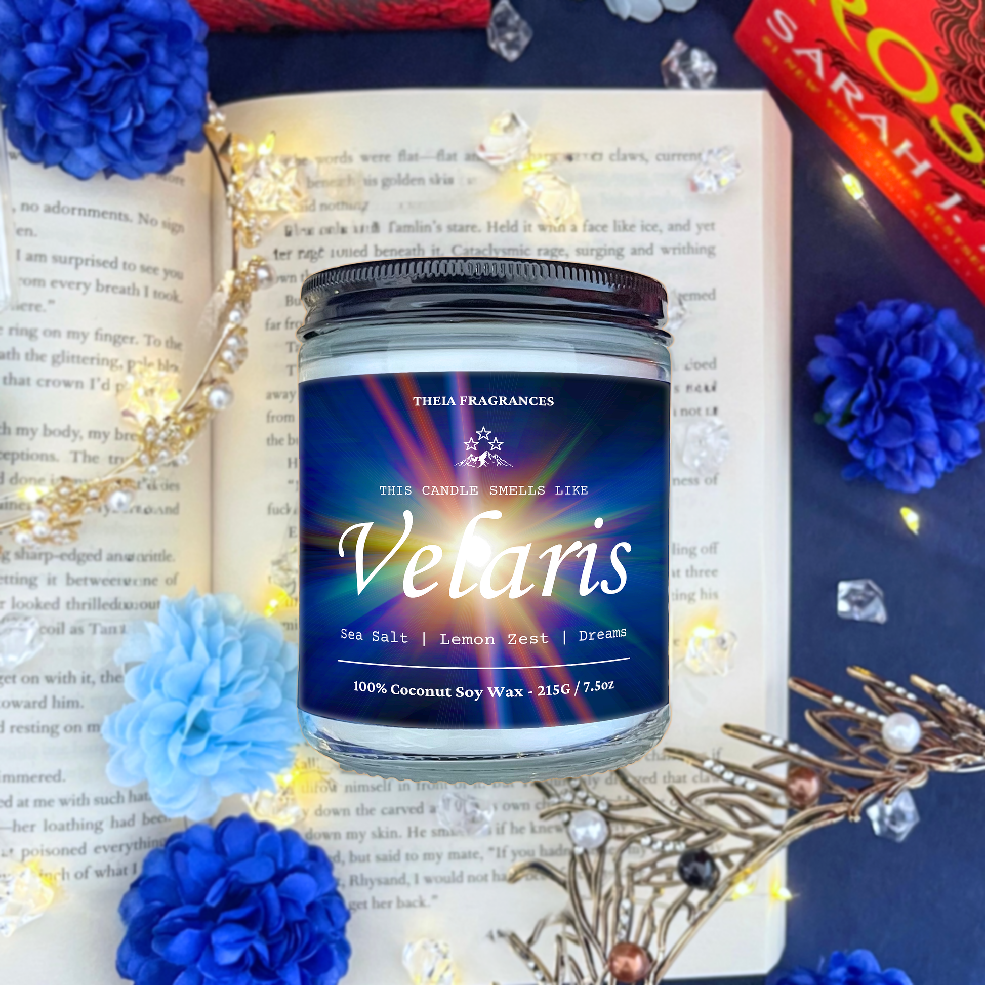 The 'Velaris' coconut soy wax candle by Theia Fragrances is demonstrated in the 215G glass jar with a black lid. The label features a burst of starlight. It is styled on an open book with decorative crystals and flowers.